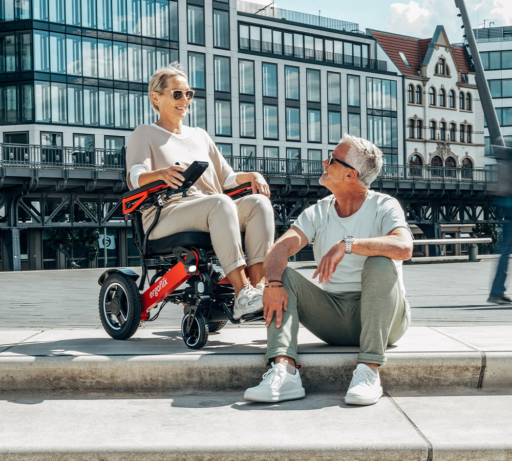 Eine ältere Dame sitzt gut gelaunt in einem roten ergoflix® LX Rollstuhl und unterhält sich mit einem Herrn, der im freien auf einer Stufe sitzt und die Sonne genießt.